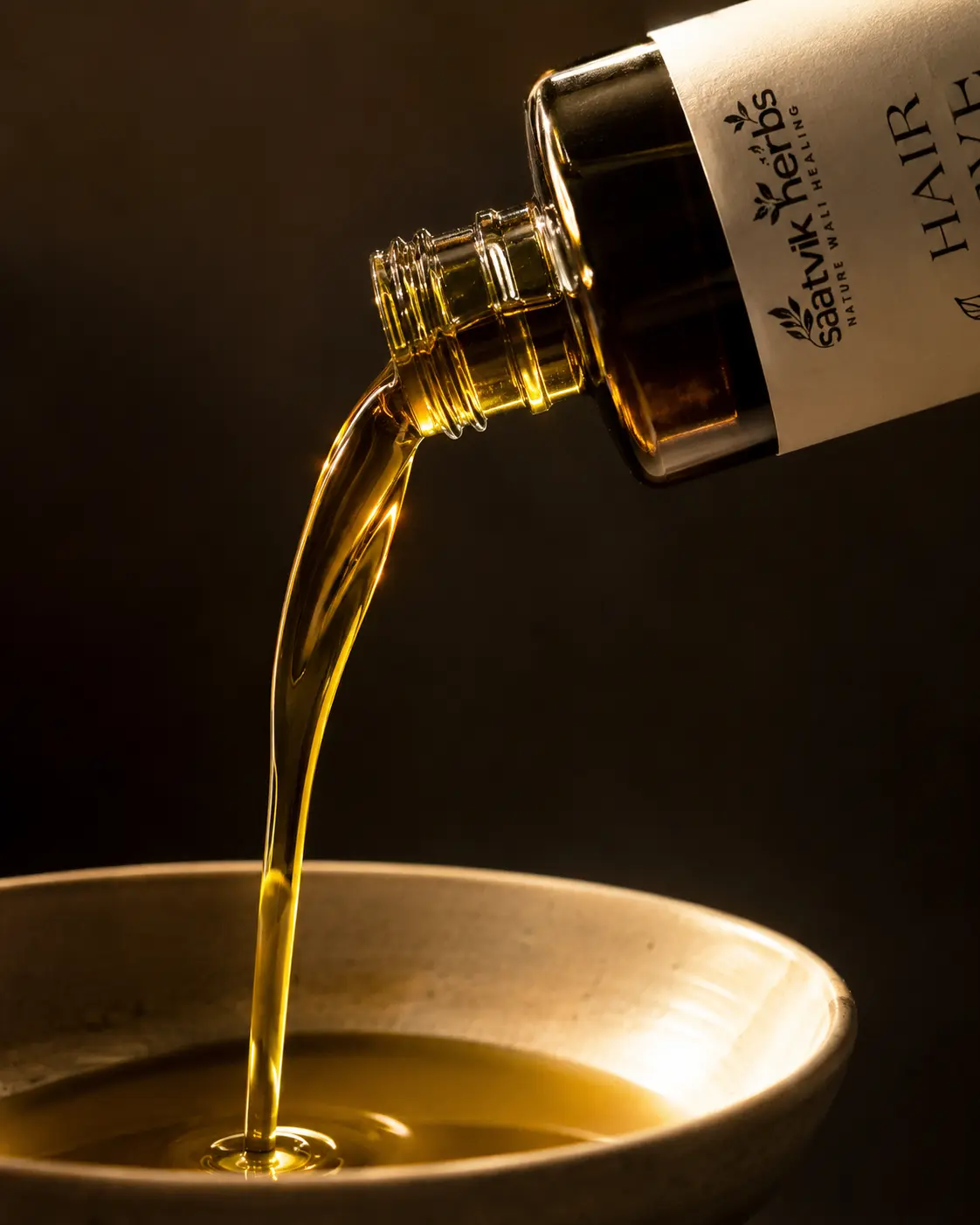 Hair Jeevan oil being poured from the bottle into a shallow ceramic bowl, lit from the side against a dark background